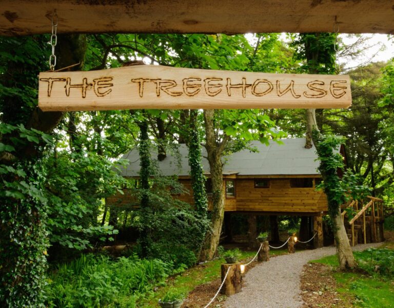 A wooden sign reading "The Treehouse" hangs in front of a large treehouse surrounded by green trees and a gravel path.