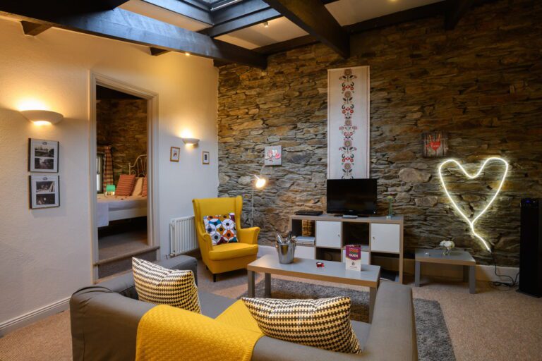 Modern living room with exposed stone wall, yellow accents, armchair, sofa, TV, and glowing heart-shaped wall light; bedroom visible through open doorway.
