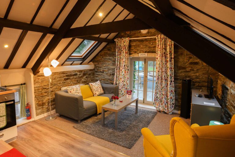 Cosy attic living room with exposed beams, stone walls, two sofas, a yellow armchair, coffee table, TV, and glass doors with floral curtains leading outside.