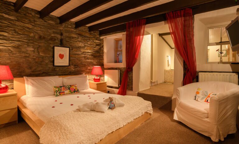 Cosy bedroom with wooden beams, stone wall, double bed, red lamps, white armchair, and red sheer curtains leading to a bathroom. Towels and rose petals are on the bed.