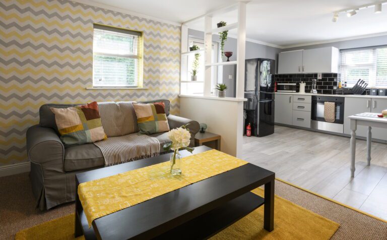 A modern open-plan living room and kitchen with patterned wallpaper, a grey sofa, a black coffee table, and a small dining area.