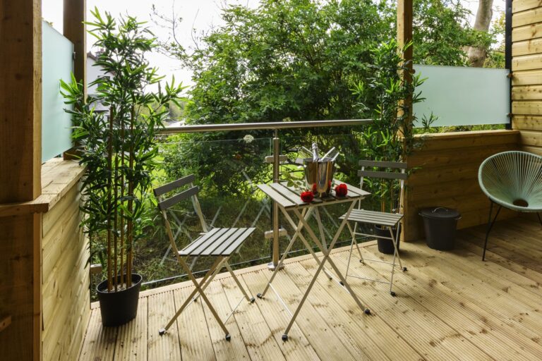 A small balcony with a metal table and two chairs, potted plants, and a champagne bucket with glasses, overlooking greenery through a glass balustrade.