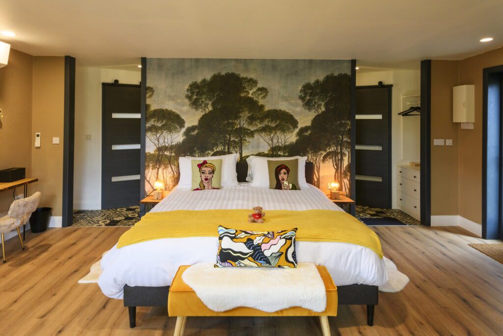 A modern bedroom with a large bed, pop art cushions, yellow throw, wooden floors, and a wall mural of trees behind the bed.