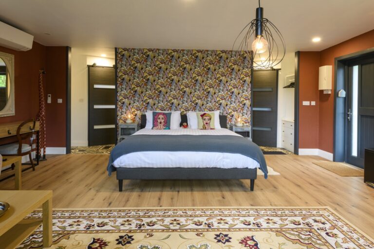 Modern bedroom with a patterned feature wall, double bed featuring illustrated pillows, wooden floor, area rug, and eclectic decor including a desk and pendant light.