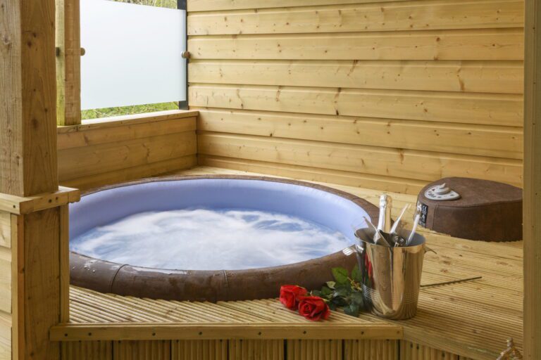 A round hot tub filled with bubbling water sits on a wooden decking. Nearby are two red roses and a champagne bucket holding a bottle and two glasses.