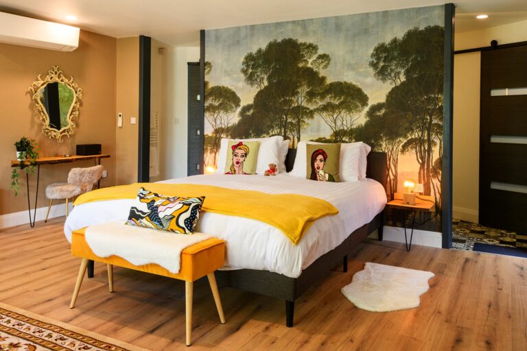 A modern bedroom with a large bed, colourful pillows and blanket, a decorative mural of trees on the wall, a mirrored dressing table, and wooden flooring.