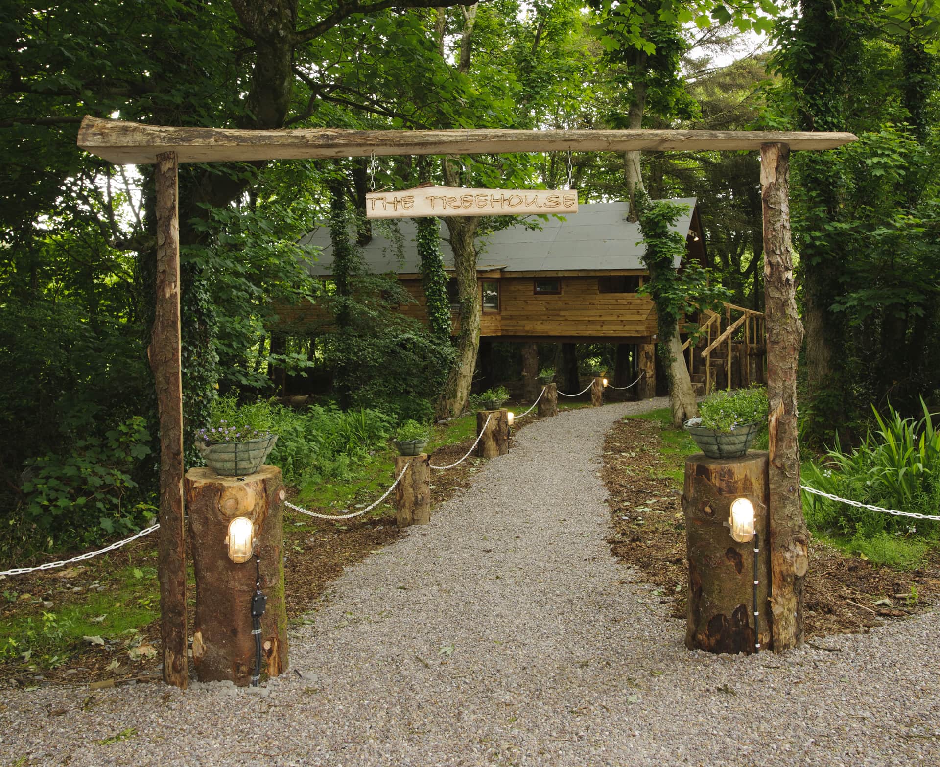 A gravel path leads to a wooden treehouse, with a rustic sign overhead reading "The Treehouse," surrounded by trees and greenery.
