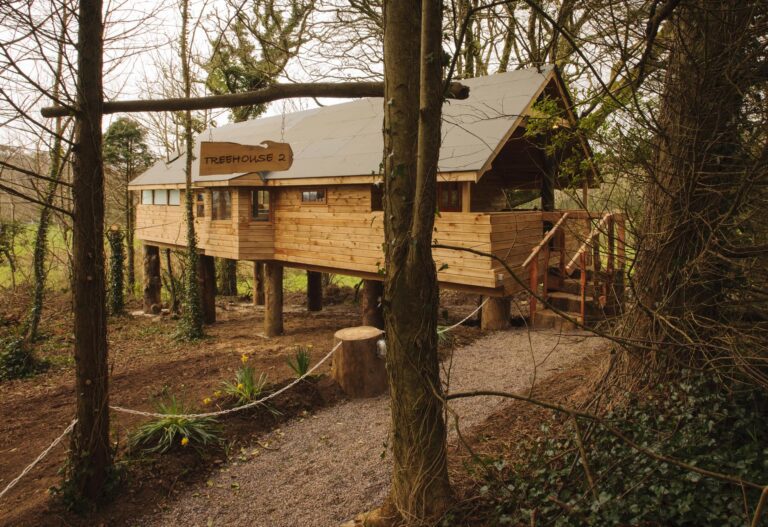 A wooden treehouse on stilts is built among tall trees, with a sign reading "Treehouse 2" above the entrance and a gravel path leading to the door.