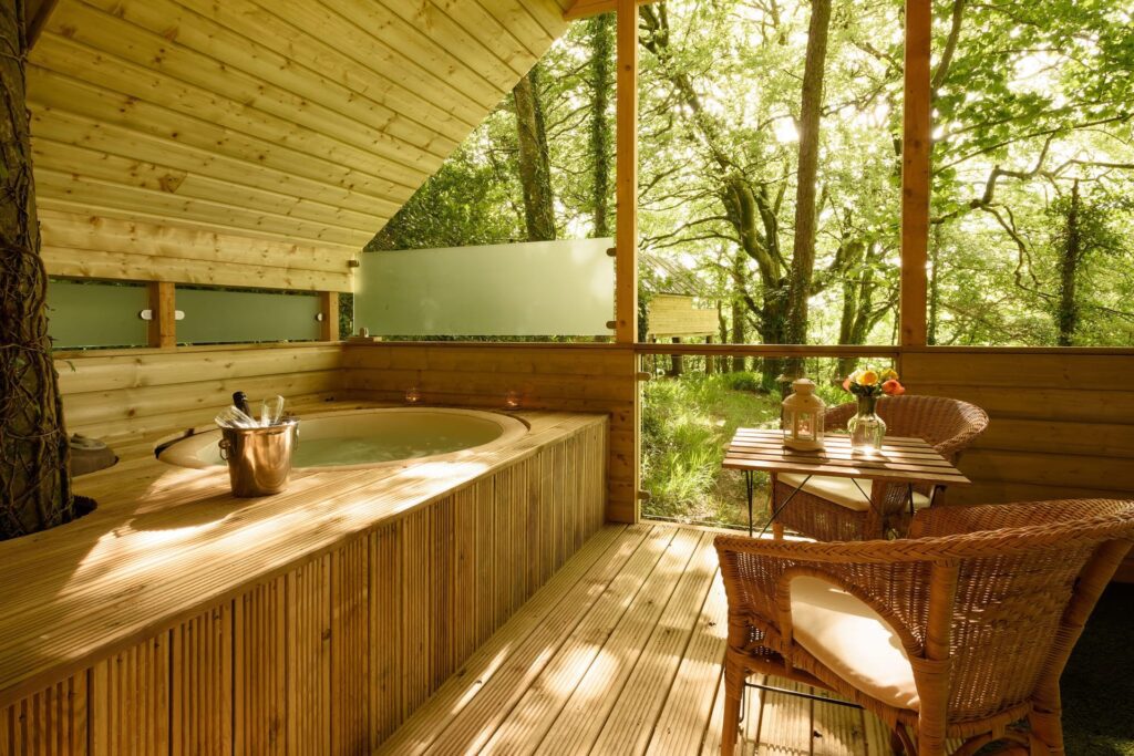 A wooden decking features a built-in round hot tub, wicker chairs, a small table with flowers, and overlooks a lush, sunlit forest through open walls.