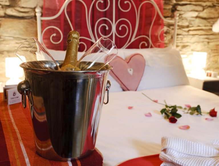 Gift Vouchers 1 A bucket with a champagne bottle and two glasses is placed on a bed, which is decorated with rose petals and a heart-shaped cushion.