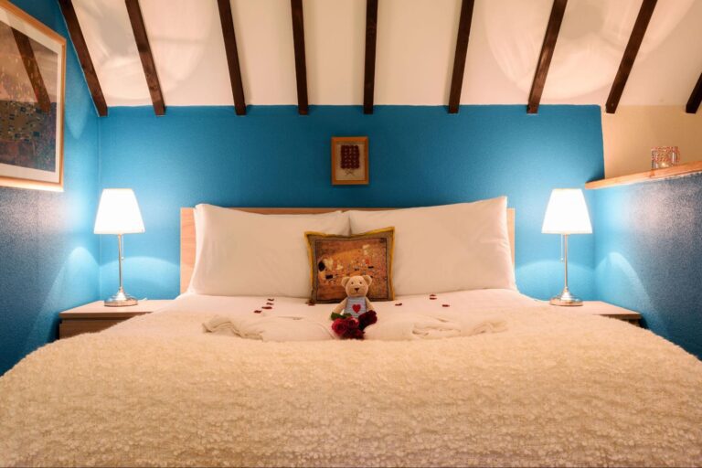 A neatly made bed with white bedding, two white pillows, a decorative cushion, a teddy bear, rose petals, and two lit lamps on bedside tables against a blue feature wall.