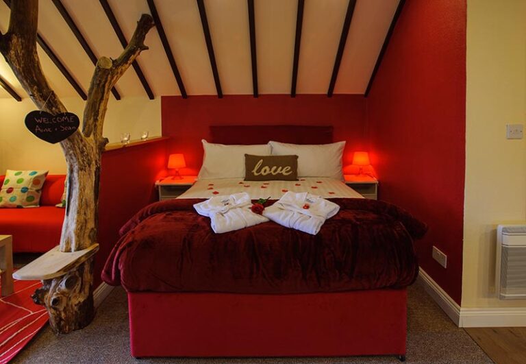 Gift Vouchers 2 A cosy bedroom with a red bed, white pillows, and a "love" cushion, decorated with rose petals and towels; warm lighting and a tree trunk with a "Welcome" sign beside the bed.