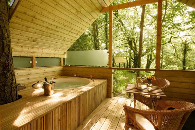 A wooden outdoor decking with a hot tub, wicker chairs, a small table, and a forest view through large open windows.