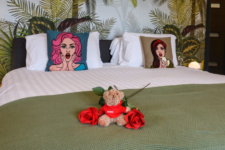 Gift Vouchers 3 A neatly made bed with two cartoon-themed pillows, a green blanket, a teddy bear holding a red heart, and two red roses placed on top.