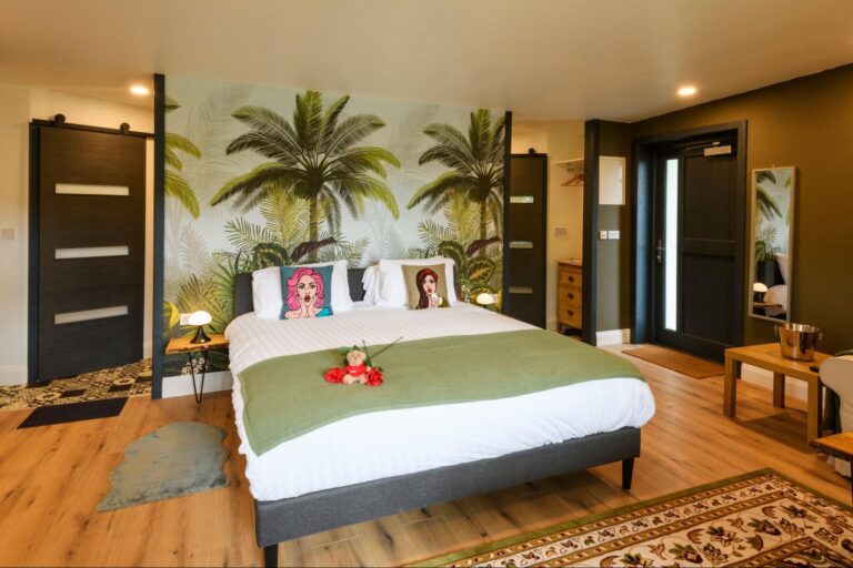 About Us 3 A modern bedroom with a double bed, tropical palm tree wallpaper, two illustrated pillows, and green décor accents. A small table with a vase of red flowers is on the bed.