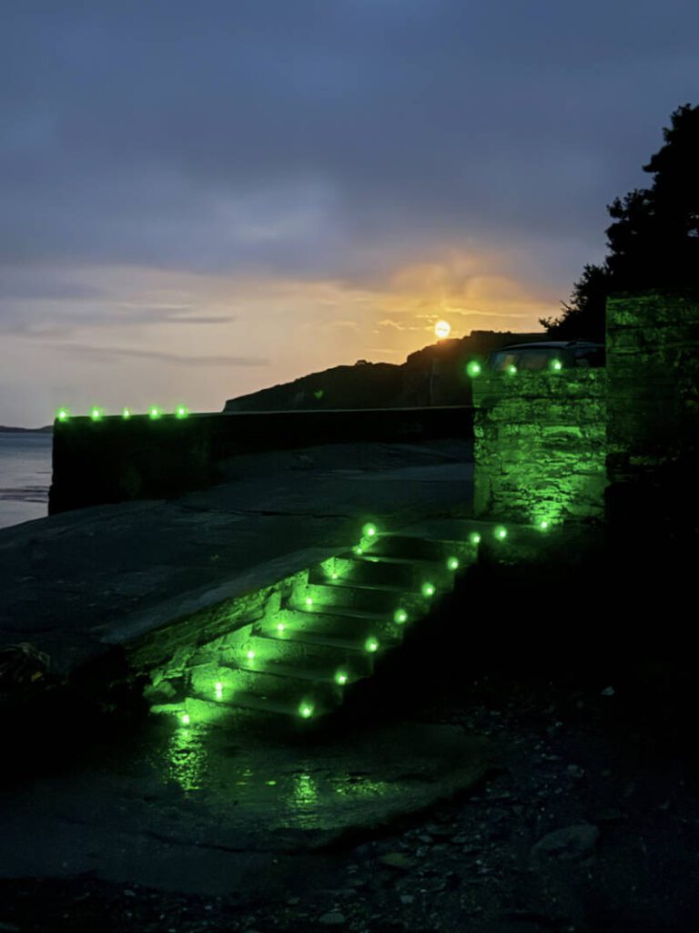 Couple Experiences 3 Stone steps with green LED lights descend towards a waterfront at dusk, with a sunset visible behind a hill in the background.