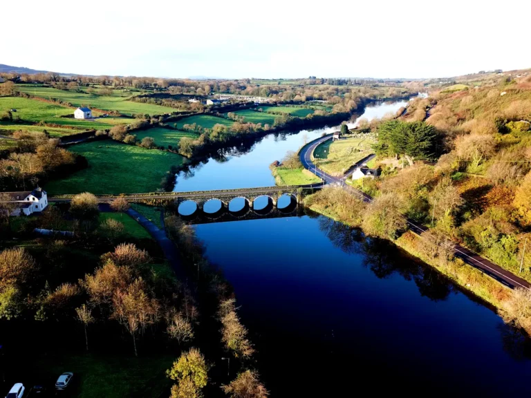 Aerial view of a stone bridge with circular arches crossing over a calm river, surrounded by green fields, trees, and a winding road.
