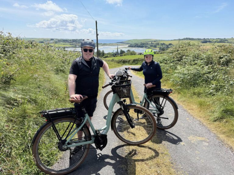 Couple Experiences 2 Two people wearing helmets stand with electric bikes on a rural path, surrounded by greenery with a lake and hills in the background under a blue sky.