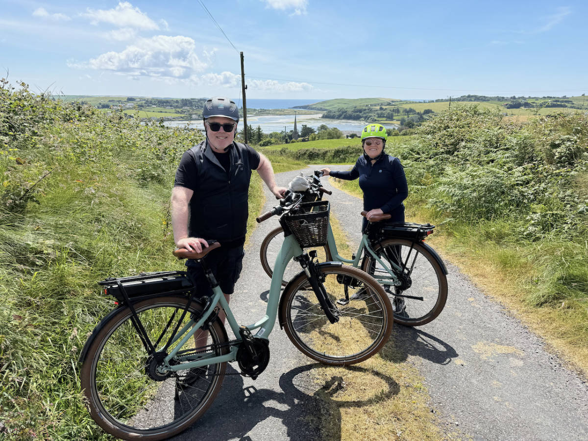 Two people wearing helmets stand with electric bikes on a rural path, surrounded by greenery with a lake and hills in the background under a blue sky.