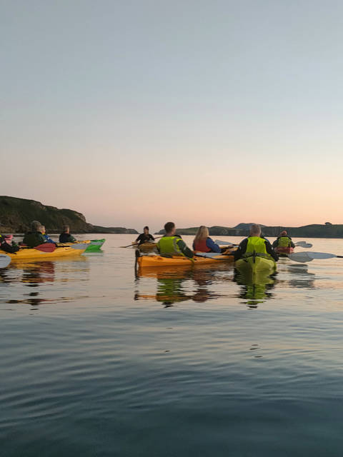 A group of people in kayaks paddle on calm water at sunset, with low hills visible in the background.