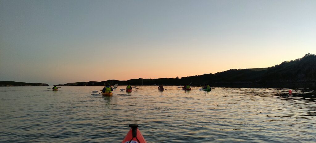 A group of kayakers paddles on calm water at sunset, with hills silhouetted in the background and a kayak's bow visible in the foreground.