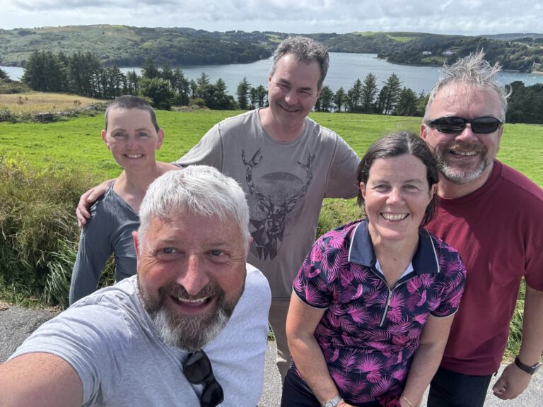 Couple Experiences 5 Five adults smiling and posing for a selfie outdoors, with lush green hills, trees, and a body of water in the background under a partly cloudy sky.