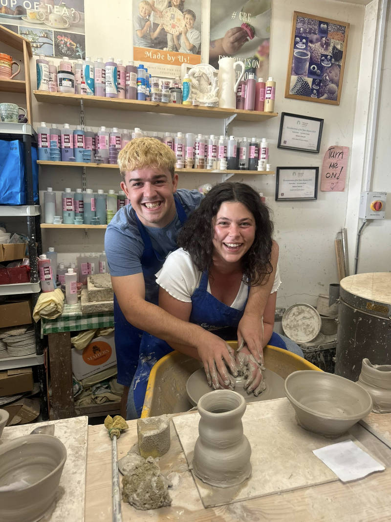 Two people wearing aprons smile while making pottery on a wheel in a ceramics studio. Finished clay pots and jars are on the table in front of them.