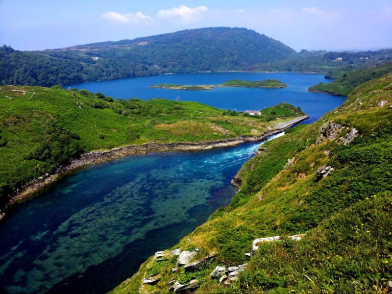 A lush green landscape with a winding river flowing into a blue lake, surrounded by hills and small islands under a partly cloudy sky.