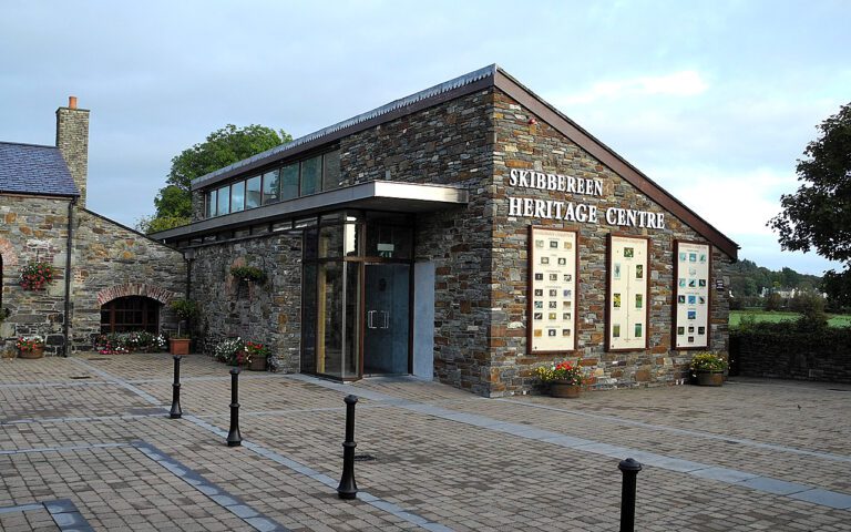 Modern stone building with large windows and flowerpots, labelled "Skibbereen Heritage Centre," set in a paved courtyard with bollards and landscaped greenery.